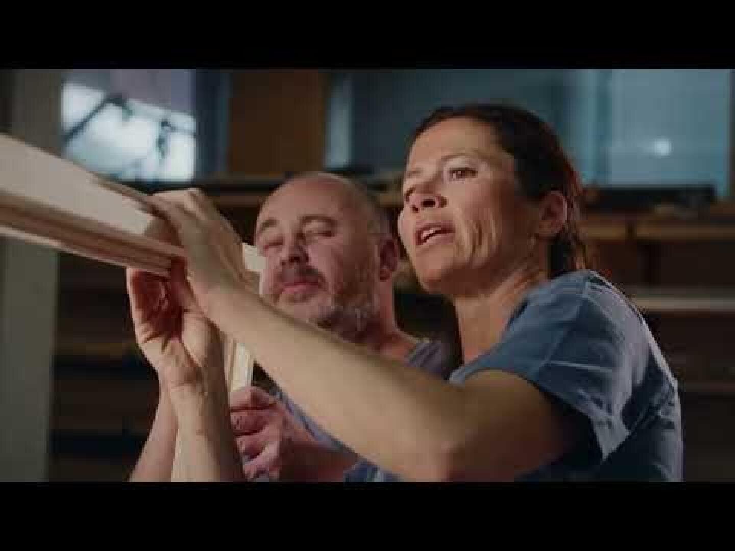 Eine Frau und ein Mann arbeiten gemeinsam an der Inspektion eines Holzbretts in einer Werkstatt, beide konzentriert auf ihre Aufgabe. Im Hintergrund sind Regale und Holzmaterialien zu sehen.