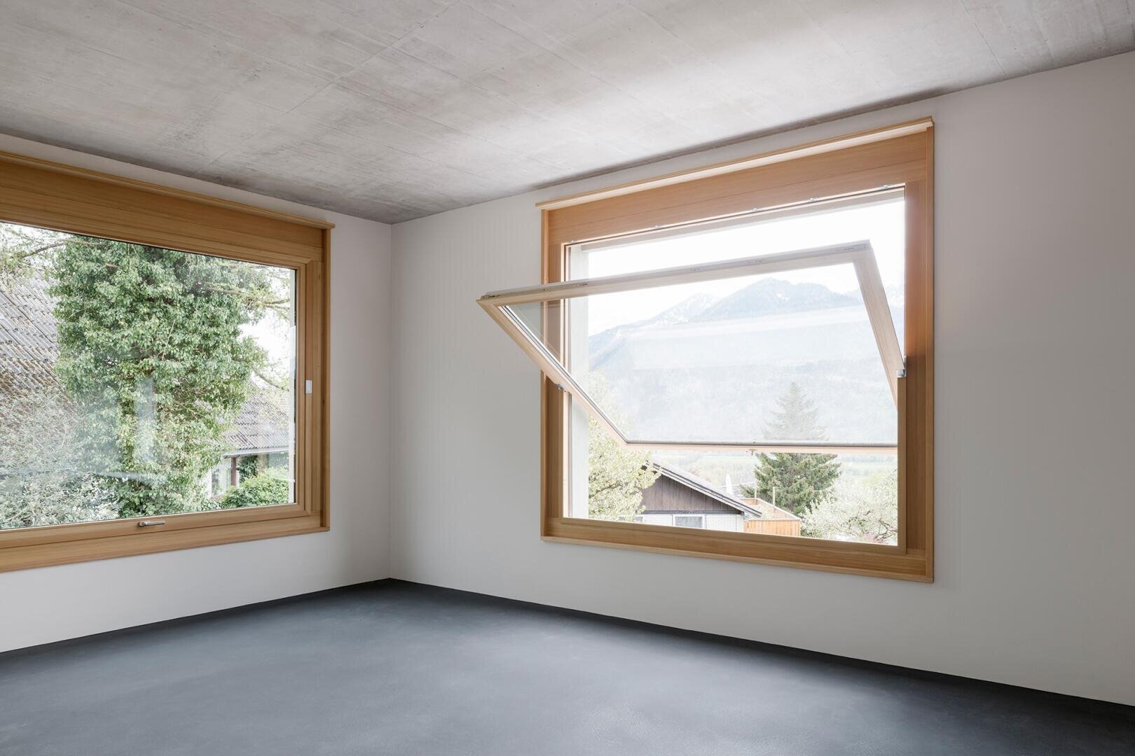 Ein moderner, leerer Raum mit großen, holzgerahmten Fenstern. Ein Fenster ist nach oben geöffnet und gibt den Blick frei auf die Berge, Bäume und Dächer draußen. Der Boden ist dunkel und die Decke ist aus Beton.