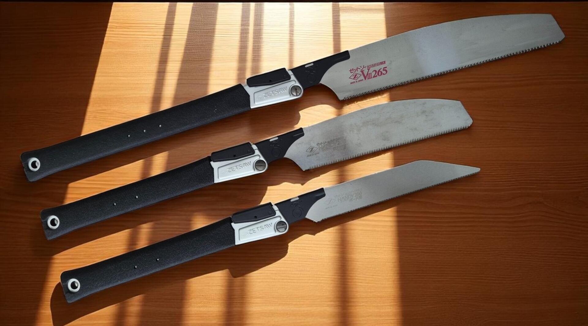 Drei japanische Sägen mit schwarzen Griffen und silbernen Blättern liegen nebeneinander auf einer hölzernen Fläche, und das Sonnenlicht wirft gestreifte Schatten auf sie.