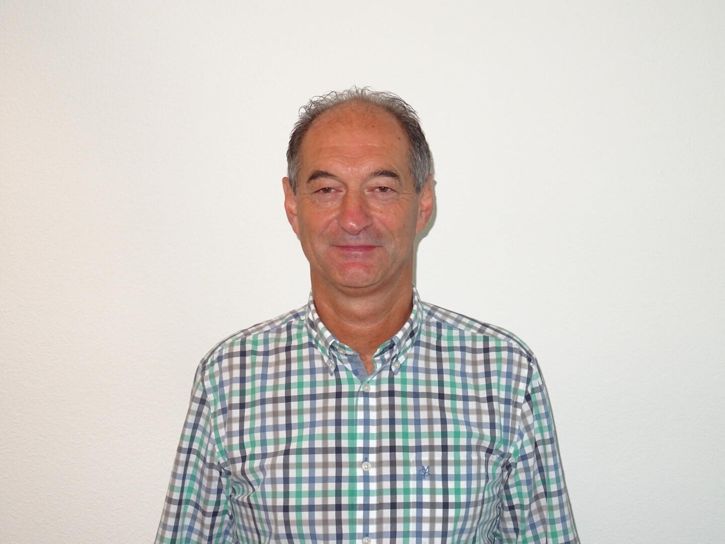 Un homme d'âge moyen aux cheveux gris courts, vêtu d'une chemise à carreaux verts, bleus et blancs, debout contre un mur blanc uni et souriant légèrement.