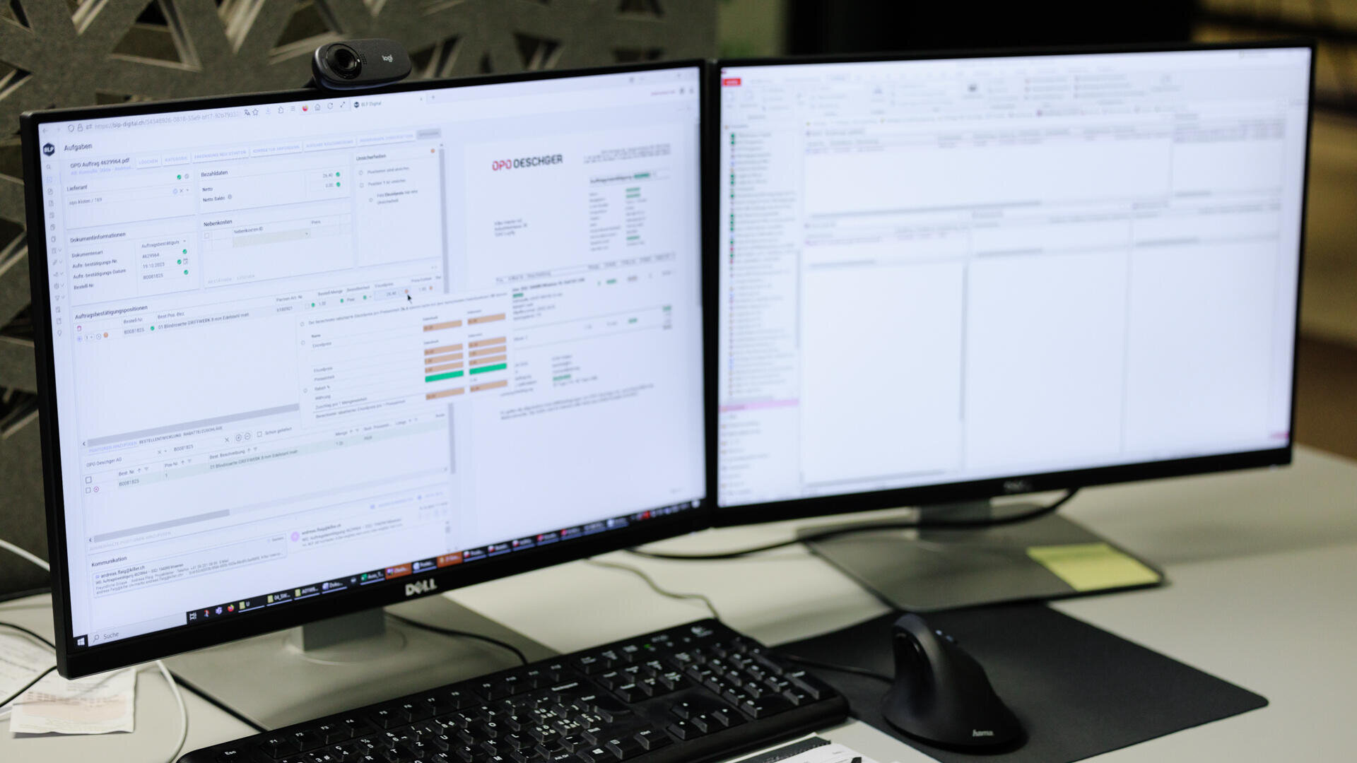 Zwei Computermonitore auf einem Schreibtisch zeigen verschiedene Softwareanwendungen und Daten an. Vor den Bildschirmen befinden sich eine schwarze Tastatur und eine Maus, auf dem linken Monitor ist eine Webcam angebracht.