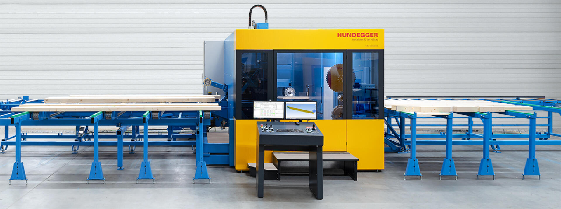 Eine gelbe Hundegger-Industrieholzbearbeitungsmaschine steht in einer Fabrik, umgeben von blauen Metallrahmen und Fördersystemen, mit Holzbrettern auf den Tischen und einem Steuerstand mit Monitoren davor.