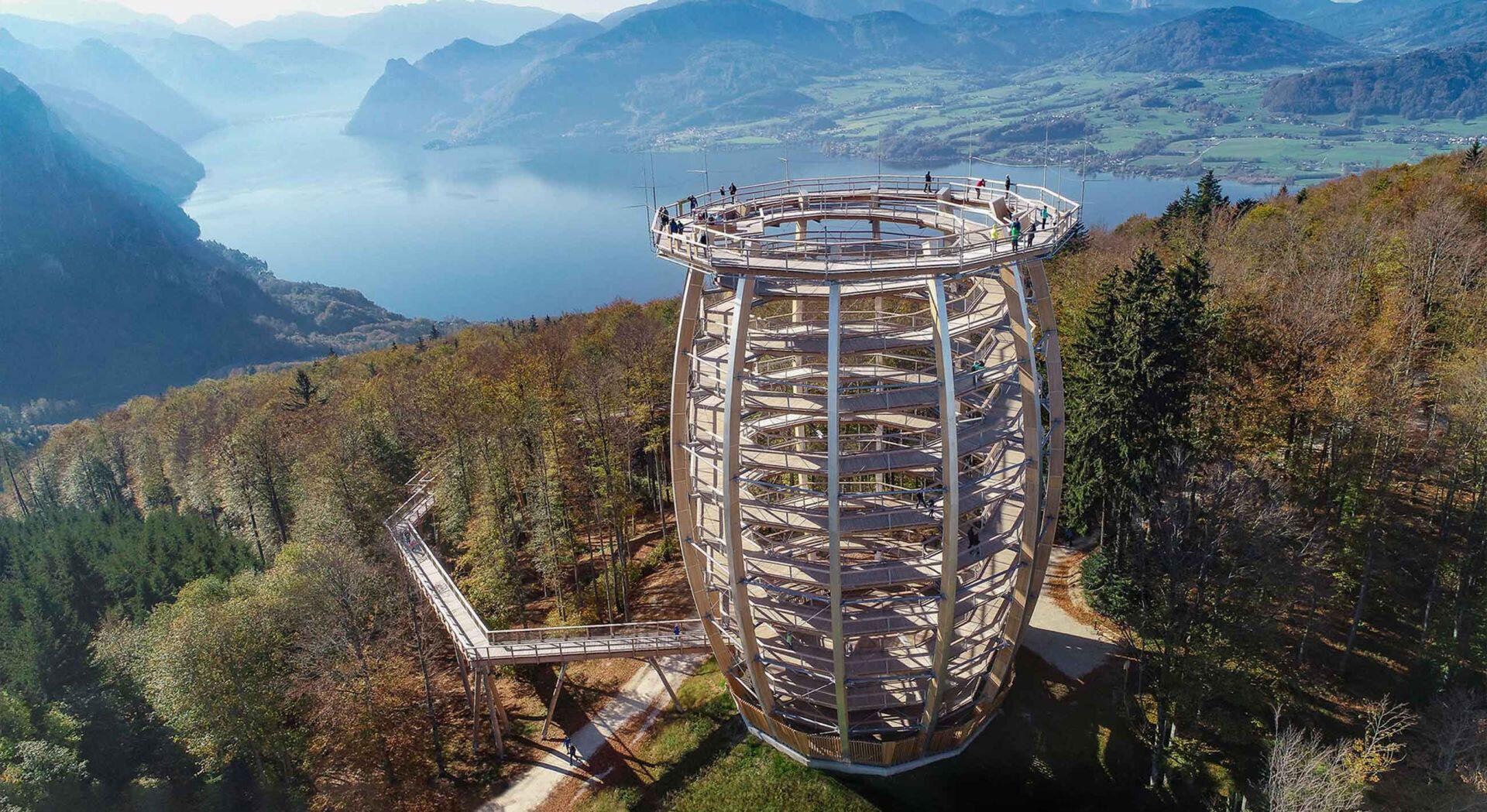 Luftaufnahme eines hohen, spiralförmigen hölzernen Aussichtsturms, der von Bäumen umgeben ist und auf dem Menschen spazieren gehen. Im Hintergrund sind Berge, ein großer See und eine malerische grüne Landschaft unter blauem Himmel zu sehen.