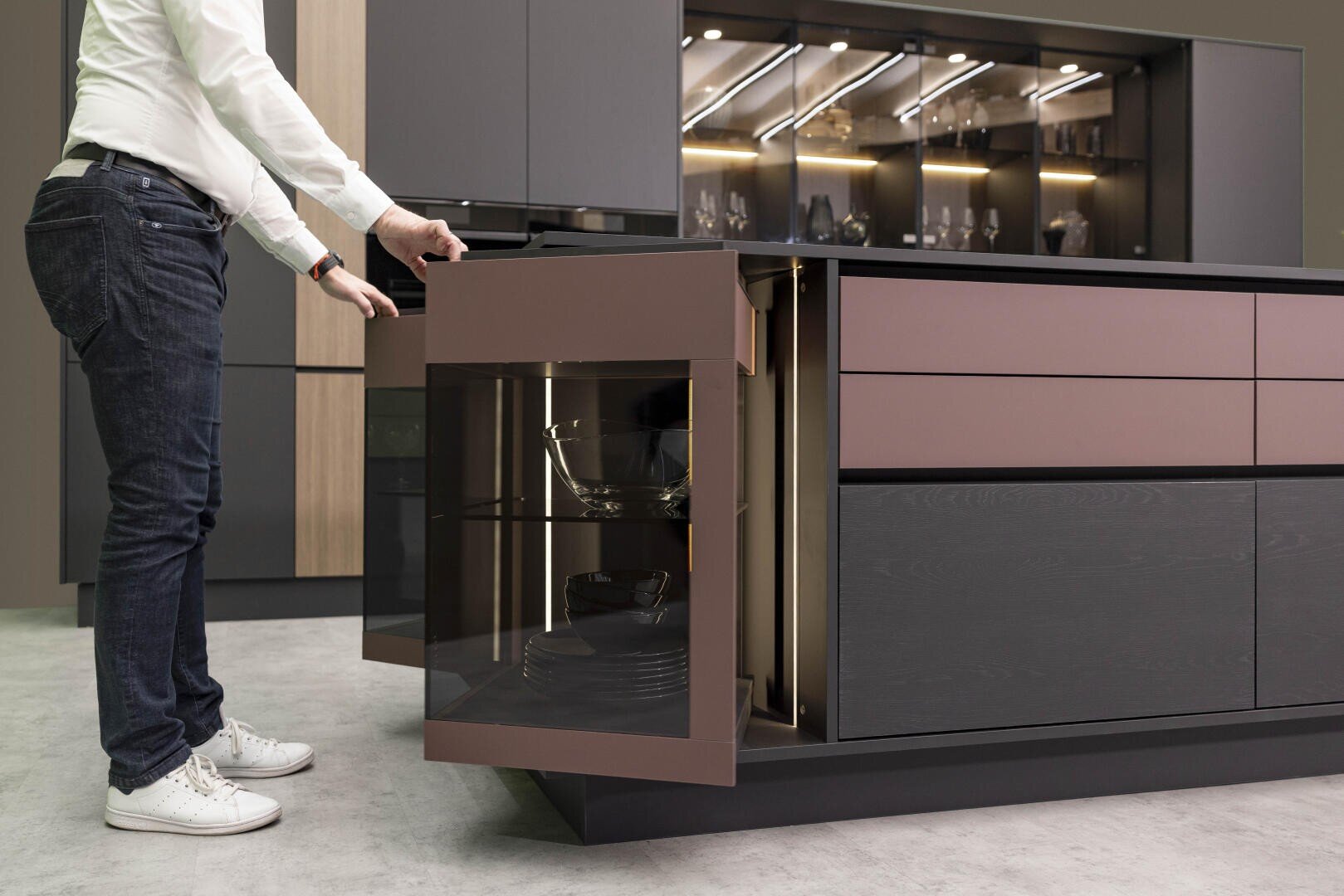 Une personne ouvre une vitrine d'angle avec une façade en verre dans une cuisine moderne avec des armoires sobres, marron foncé et noires, et laisse entrevoir des étagères avec des bols et de la vaisselle à l'intérieur.