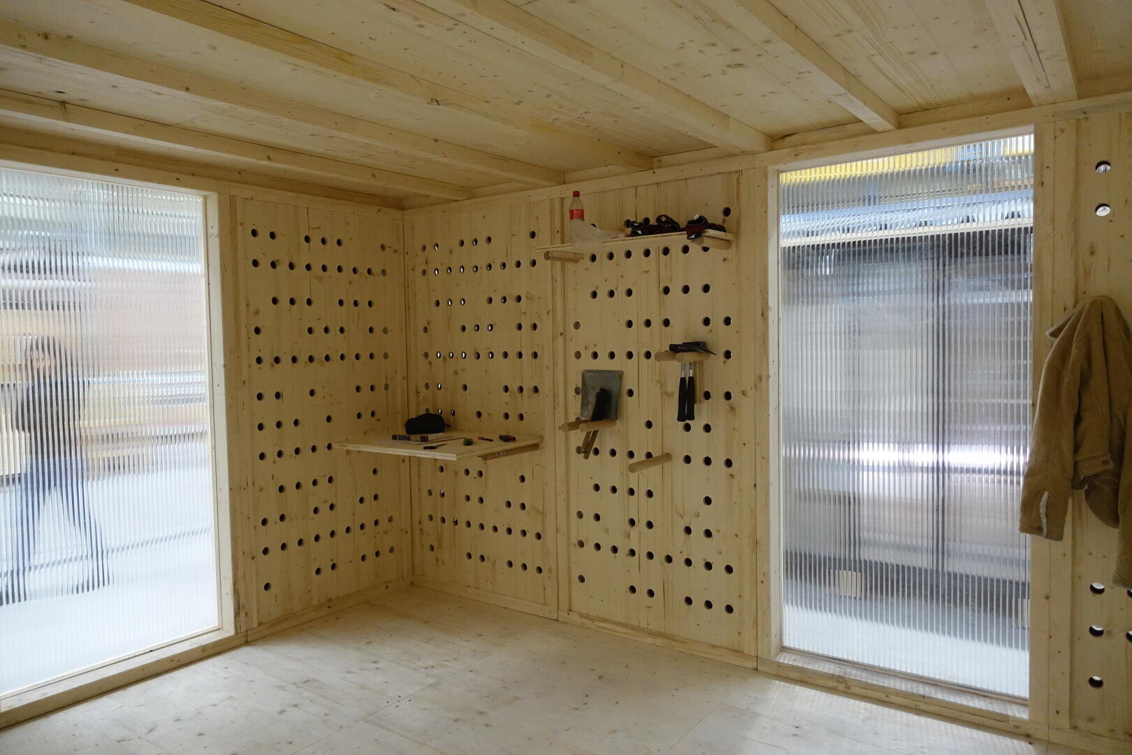 Holzzimmer mit zwei großen, durchsichtigen Fenstern, Wänden mit Stecktafeln, an denen Werkzeuge hängen, ein Mantel an einem Haken und eine kleine Werkbank mit Werkzeugen; die Menschen vor dem Fenster sind verschwommen.