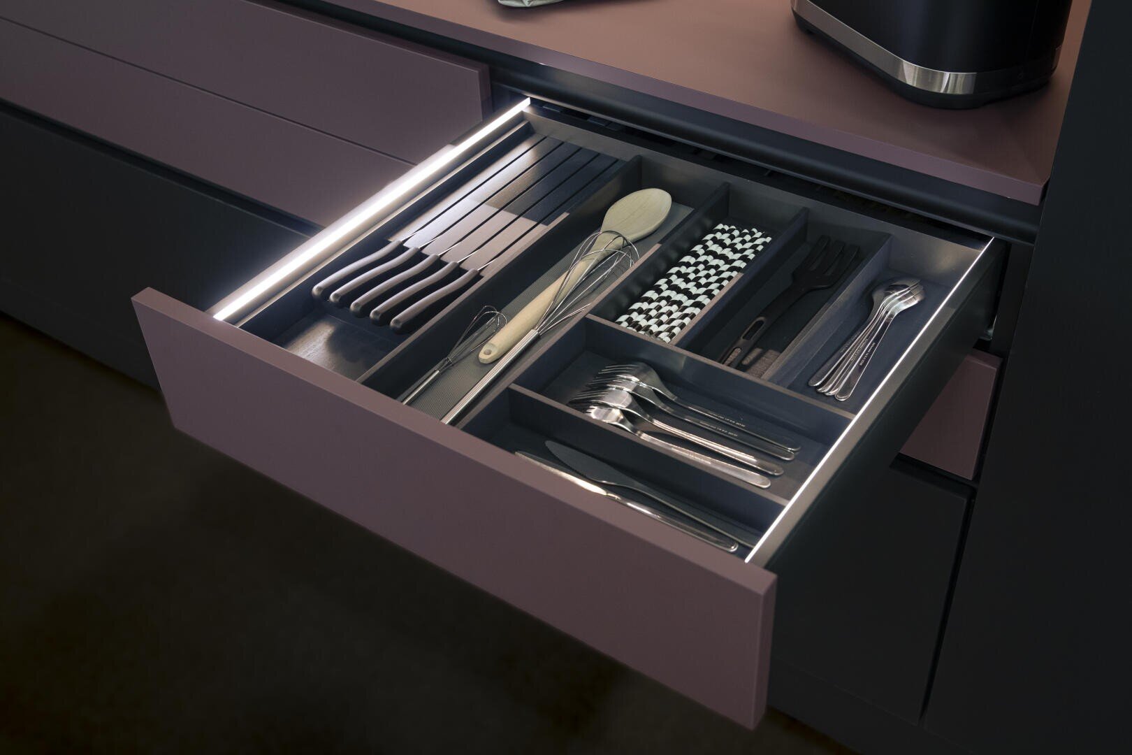 Un tiroir de cuisine ouvert avec éclairage LED intégré présente dans des compartiments séparés des couverts bien rangés, dont des couteaux, des fourchettes, des cuillères, des baguettes, des pailles, une cuillère en bois et un fouet.