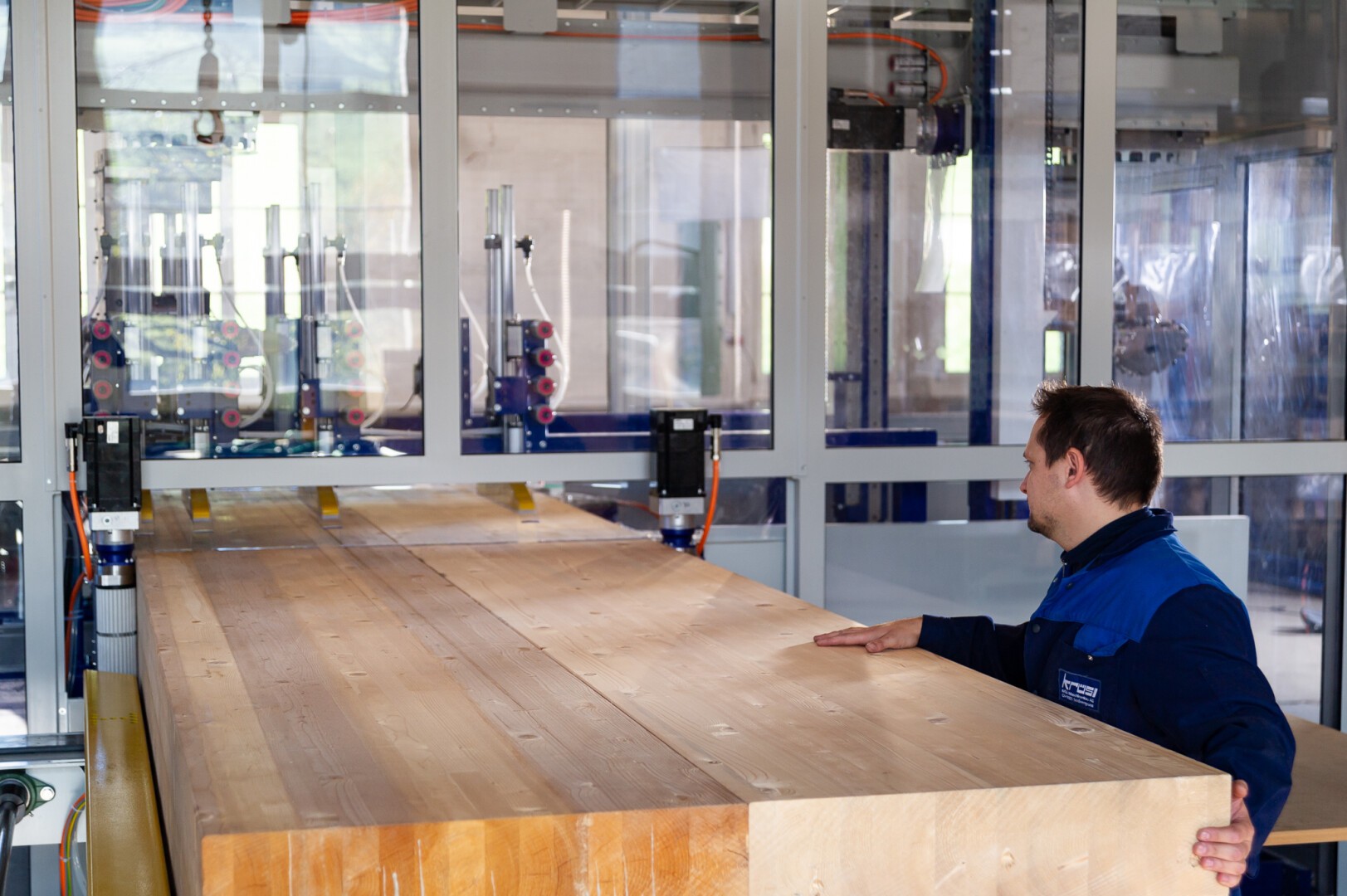 Ein Arbeiter in blauer Uniform bedient Maschinen, während er in einer industriellen Umgebung, wahrscheinlich einer Holzverarbeitungs- oder Produktionsanlage, einen großen, glatten Holzbalken handhabt.