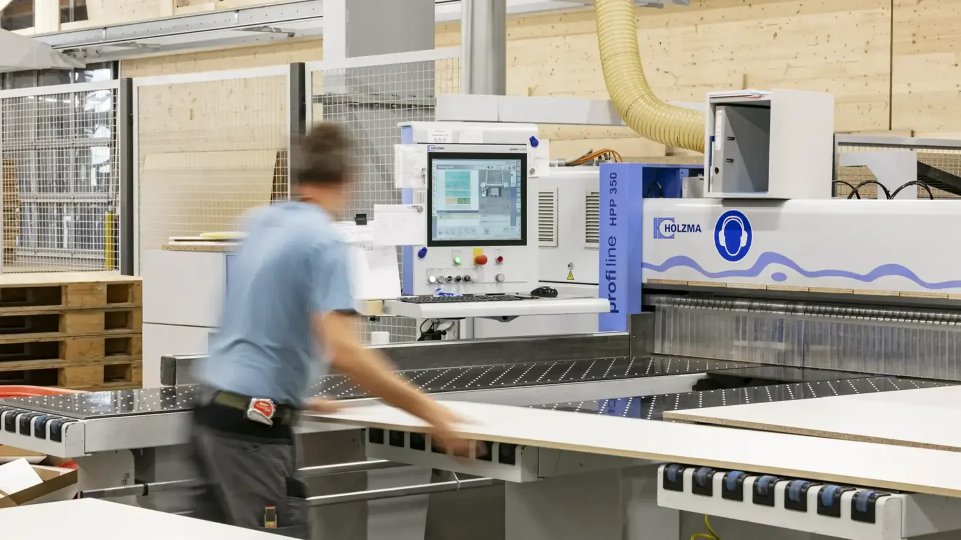 Ein Arbeiter im blauen Hemd bedient eine große industrielle CNC-Maschine, durch die er eine Materialplatte in einer Fabrikumgebung führt. Die Maschine verfügt über ein digitales Bedienfeld mit einem Bildschirm, auf dem Daten angezeigt werden.