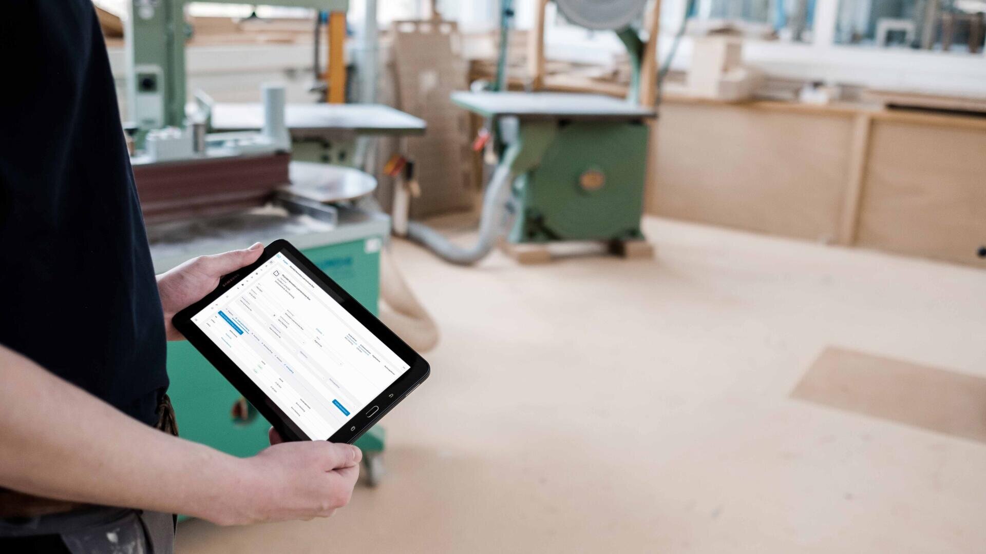 Une personne tient une tablette affichant une interface numérique dans un atelier avec des machines industrielles vertes et des surfaces en bois en arrière-plan.