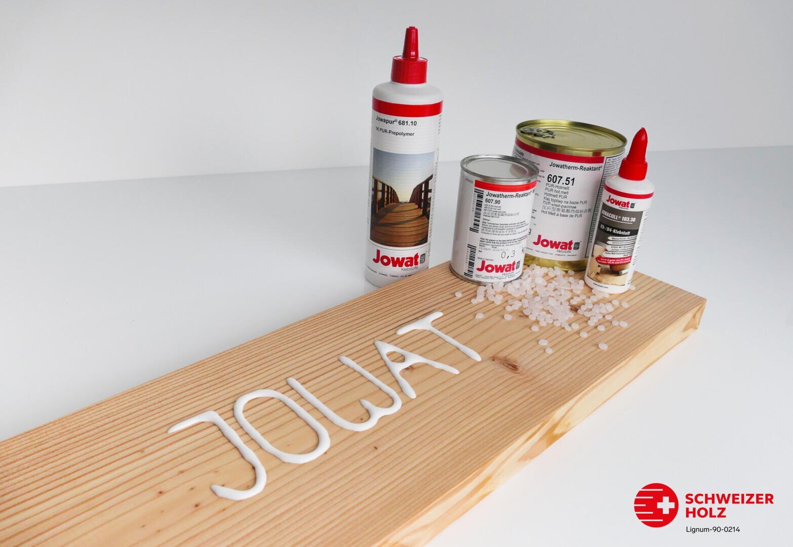 Différents produits adhésifs de Jowat sont exposés sur un panneau en bois. Le mot "JOWAT" est écrit avec de la colle sur le panneau, à côté duquel sont éparpillés des granulés de colle. Dans le coin inférieur droit, on peut voir le logo de Schweizer Holz.