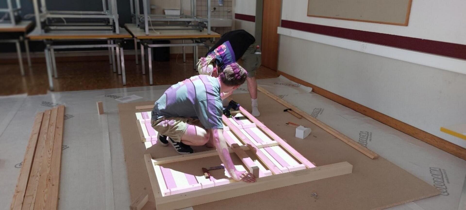 Zwei Personen bauen in einem Klassenzimmer einen Holzrahmen auf dem Boden zusammen, umgeben von Holzbrettern und Tischen, die im Hintergrund gestapelt sind. Eine Person kniet, während die andere zuschaut oder assistiert.