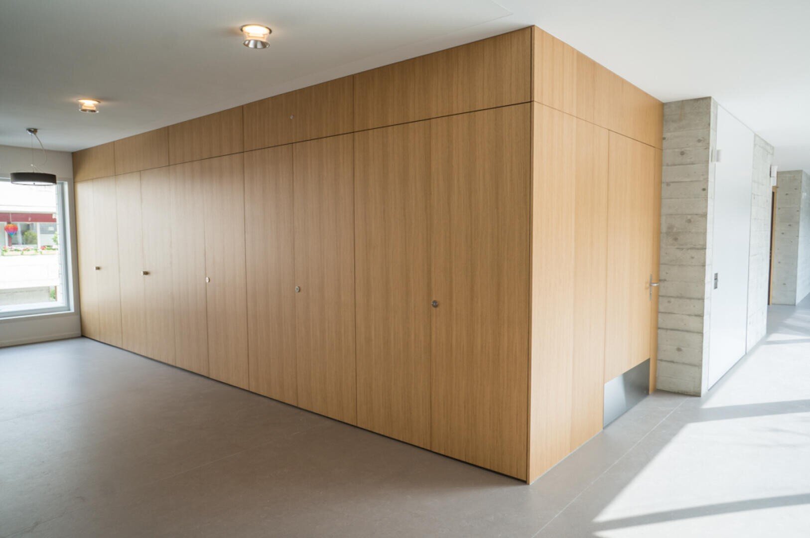 Der moderne Innenraum besticht durch eine große, hell holzgetäfelte Struktur mit flächenbündigen Türen und einer kleinen rechteckigen Haustierklappe am unteren Ende. Der Raum verfügt über Betonwände, große Fenster und einen grauen Bodenbelag.