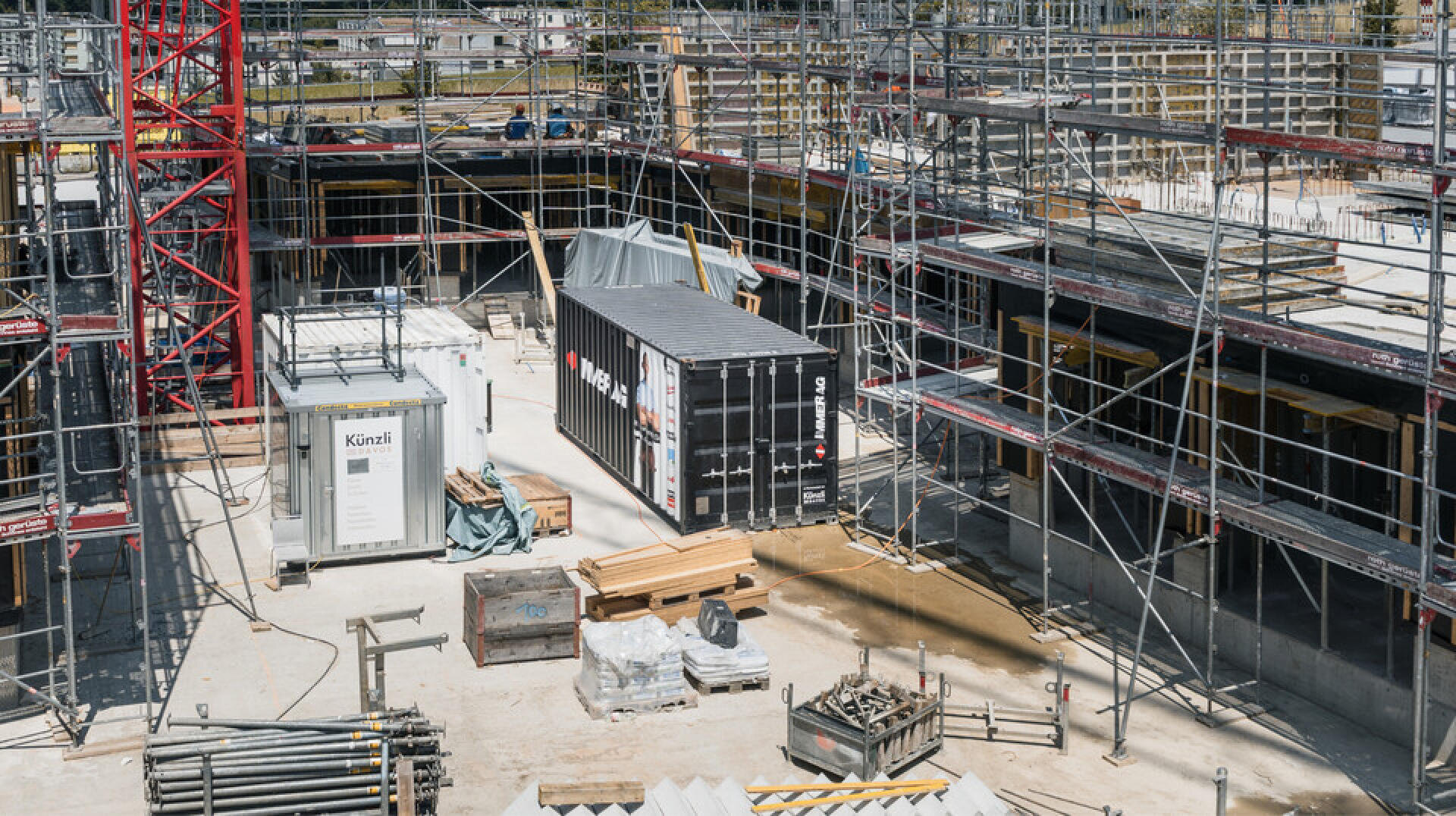 Un chantier de construction avec des échafaudages, des matériaux de construction et des conteneurs disposés autour d'une structure partiellement achevée. Des planches de bois, des tuyaux métalliques et des équipements sont éparpillés sur le sol en béton.