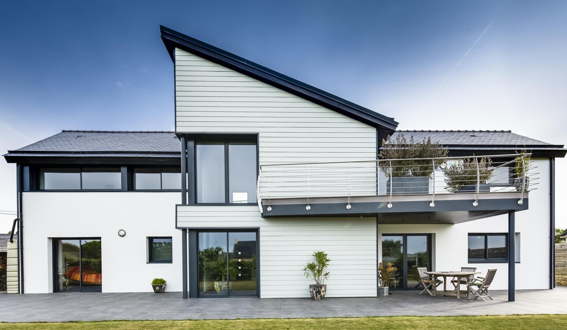 Modernes zweistöckiges Haus mit geometrischem Design, großen Fenstern, einem Balkon mit Metallgeländer und einem Sitzplatz im Freien auf einer Terrasse vor einem strahlend blauen Himmel und einer Rasenfläche.