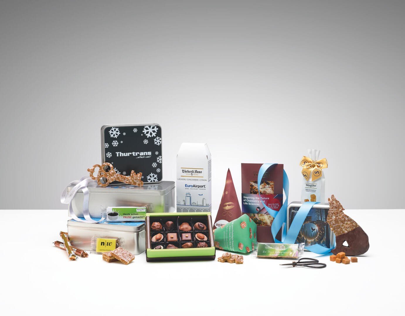 Une multitude de friandises et d'en-cas différents, de chocolats, de pâtisseries et de biscuits sont disposés sur une surface blanche avec des emballages et des étiquettes festifs. Certaines boîtes sont ouvertes pour présenter les chocolats ; des décorations et des rubans sont inclus.