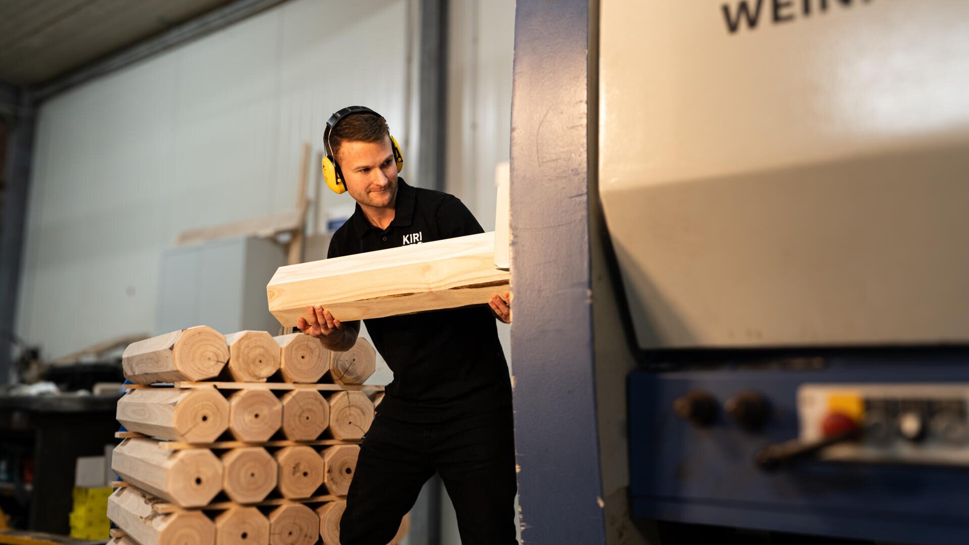 Ein Mann mit Gehörschutz legt einen Holzstamm in eine große Industriemaschine in einer Werkstatt ein. Neben ihm liegen Stapel ähnlicher Holzstämme.