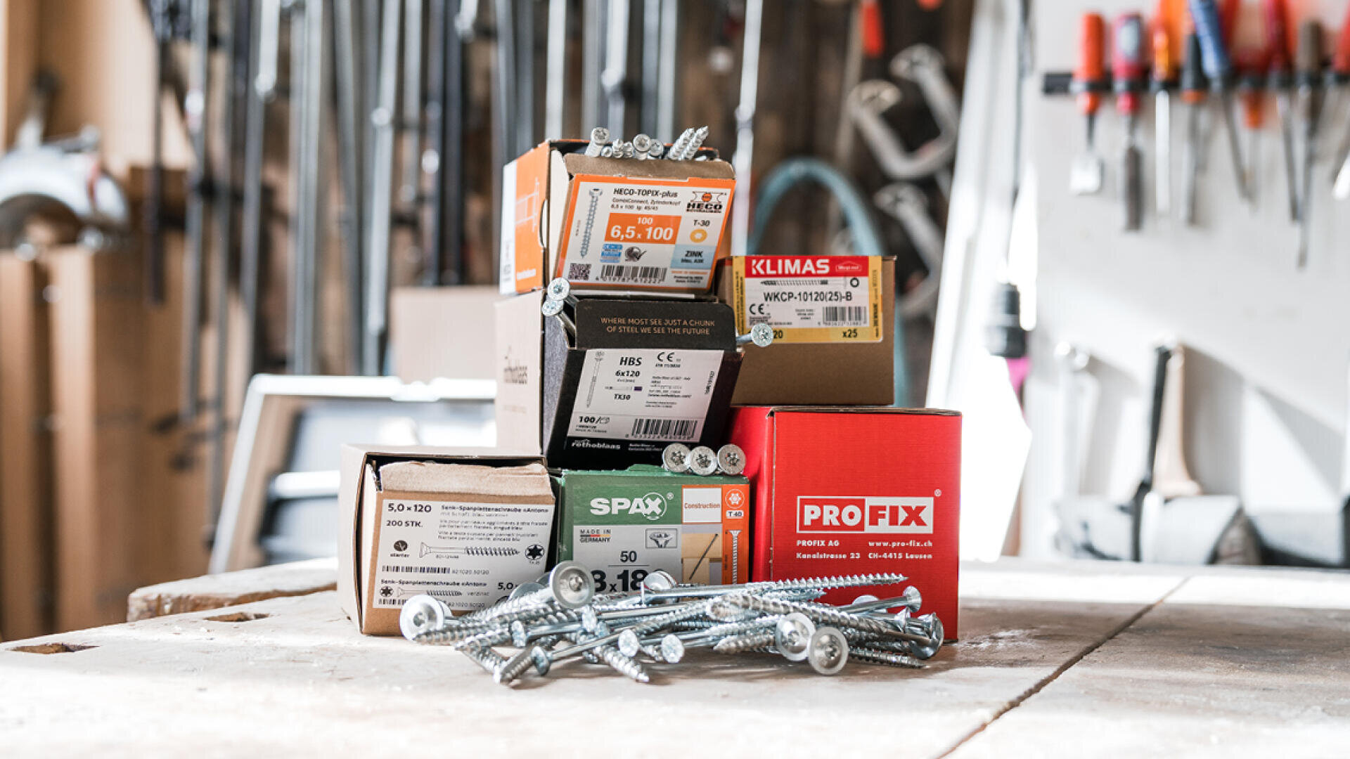 Des boîtes de vis et d'attaches diverses sont empilées sur un établi en bois dans un atelier, avec divers outils et équipements visibles dans l'arrière-plan flou. Des vis en vrac sont éparpillées au premier plan.