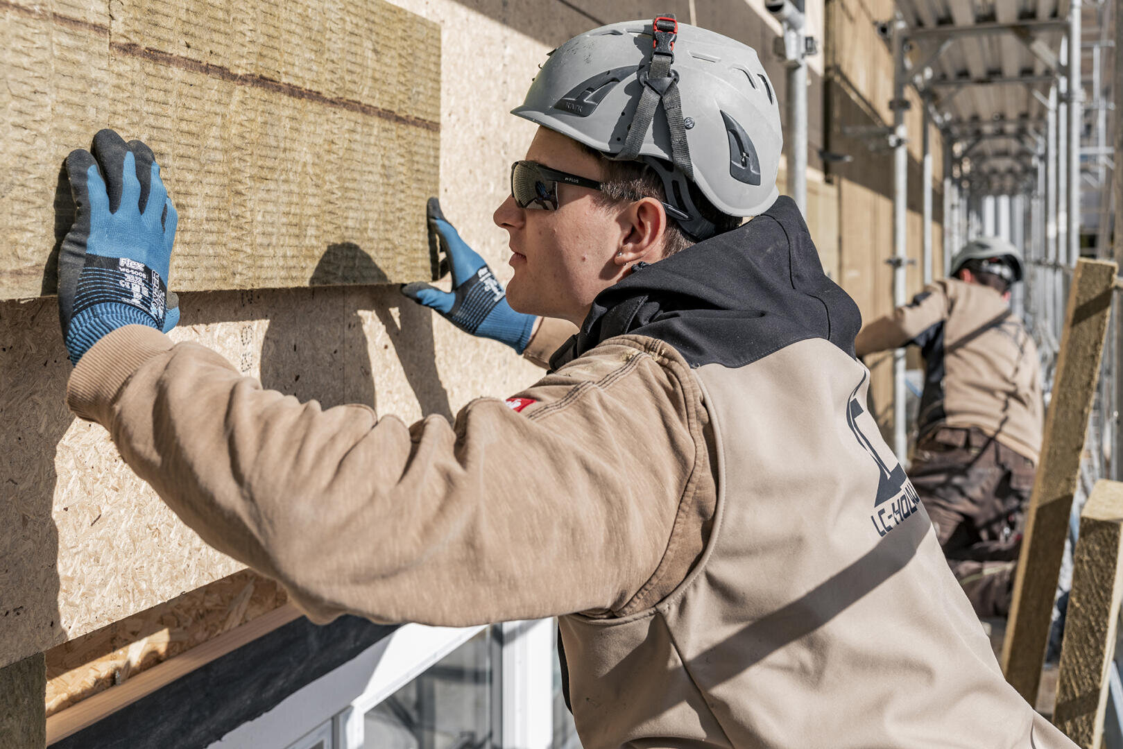 Ein Bauarbeiter mit Helm, Handschuhen und Schutzbrille bringt Dämmplatten an der Außenwand eines Gebäudes an, während ein anderer Arbeiter im Hintergrund eine ähnliche Arbeit ausführt.