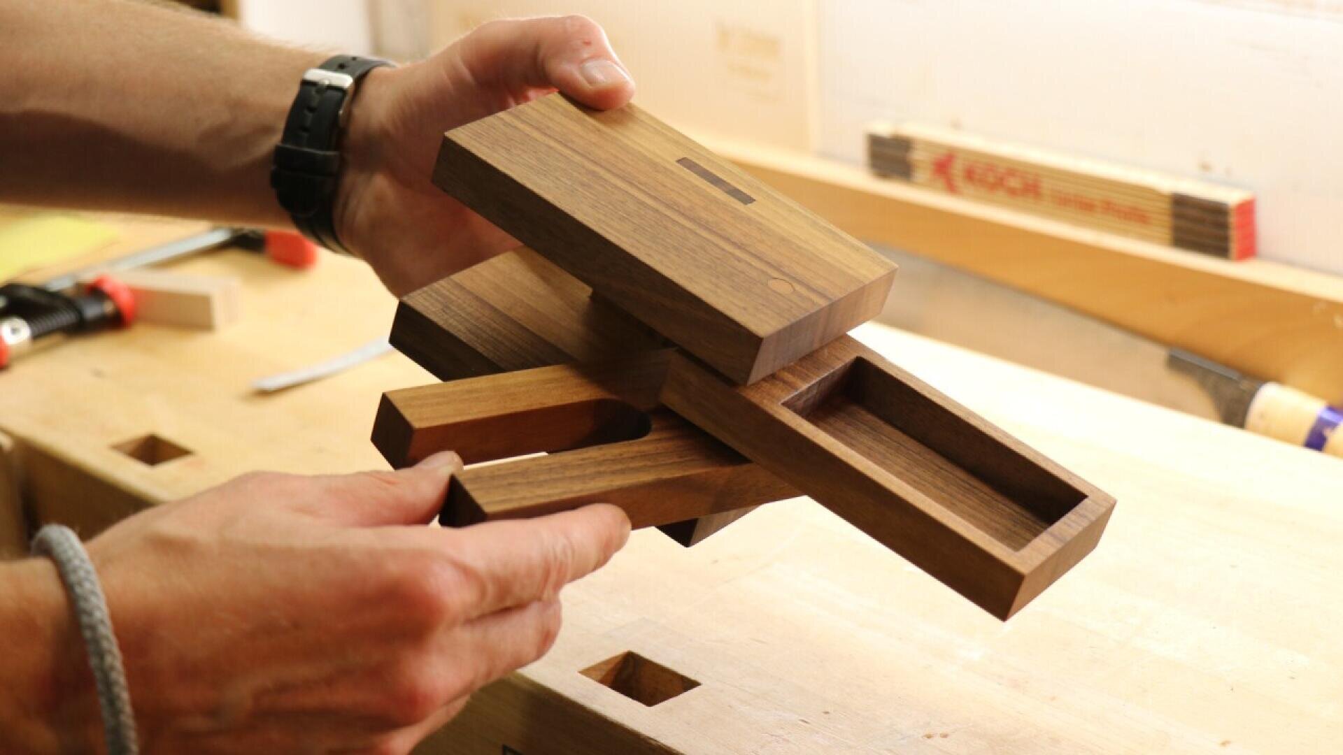 Une personne tient une boîte de puzzle en bois avec plusieurs pièces coulissantes et amovibles, démontrant son assemblage. L'arrière-plan montre un établi de menuiserie avec des outils et des blocs de bois.
