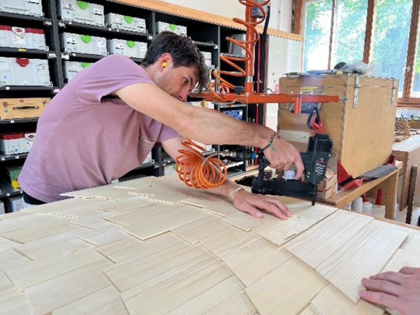 Eine Person mit Ohrstöpseln bearbeitet in einer Werkstatt dünne Holzplatten mit einem Druckluftwerkzeug. Im Hintergrund sind Regale mit geordneten Kisten und ein großes Fenster zu sehen.