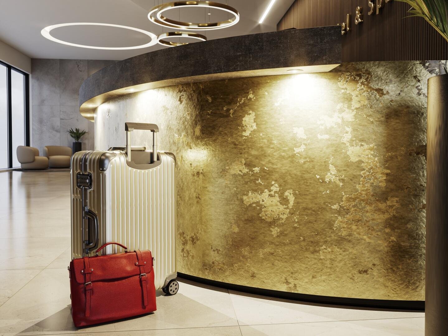 Ein silberner Koffer und ein roter Aktenkoffer stehen vor einer goldfarbenen Hotelrezeption in einer modernen, gut beleuchteten Lobby.