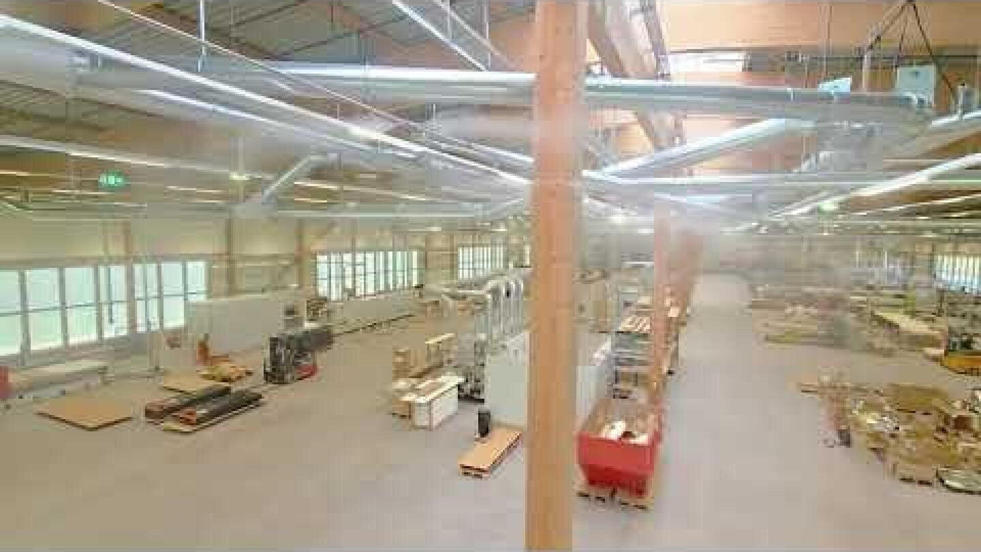 Geräumiges Fabrikinterieur mit hohen Decken, freiliegenden Kanälen und großen Fenstern. Verschiedene Maschinen, Paletten und Materialien sind in dem offenen, gut beleuchteten Bereich verteilt. Es sind keine Menschen zu sehen.