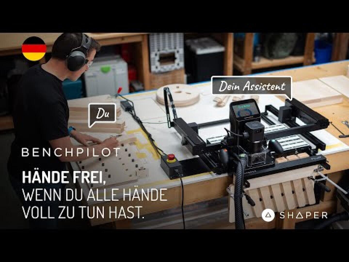 Eine Person mit Kopfhörern arbeitet an einer Holzwerkstattbank neben einer Shaper-Maschine mit der Aufschrift "Dein Assistent". Der deutsche Text darunter lautet: "Hände frei, wenn du alle Hände voll zu tun hast.".