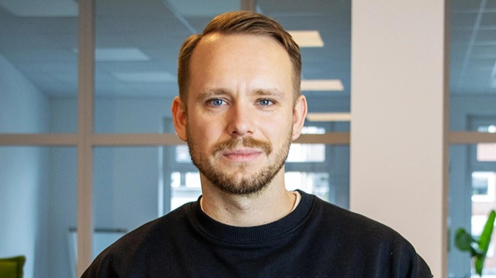 Un homme aux cheveux courts châtain clair et à la barbe taillée, portant un pull noir à col ras du cou, se tient à l'intérieur devant de grandes fenêtres avec une expression neutre.
