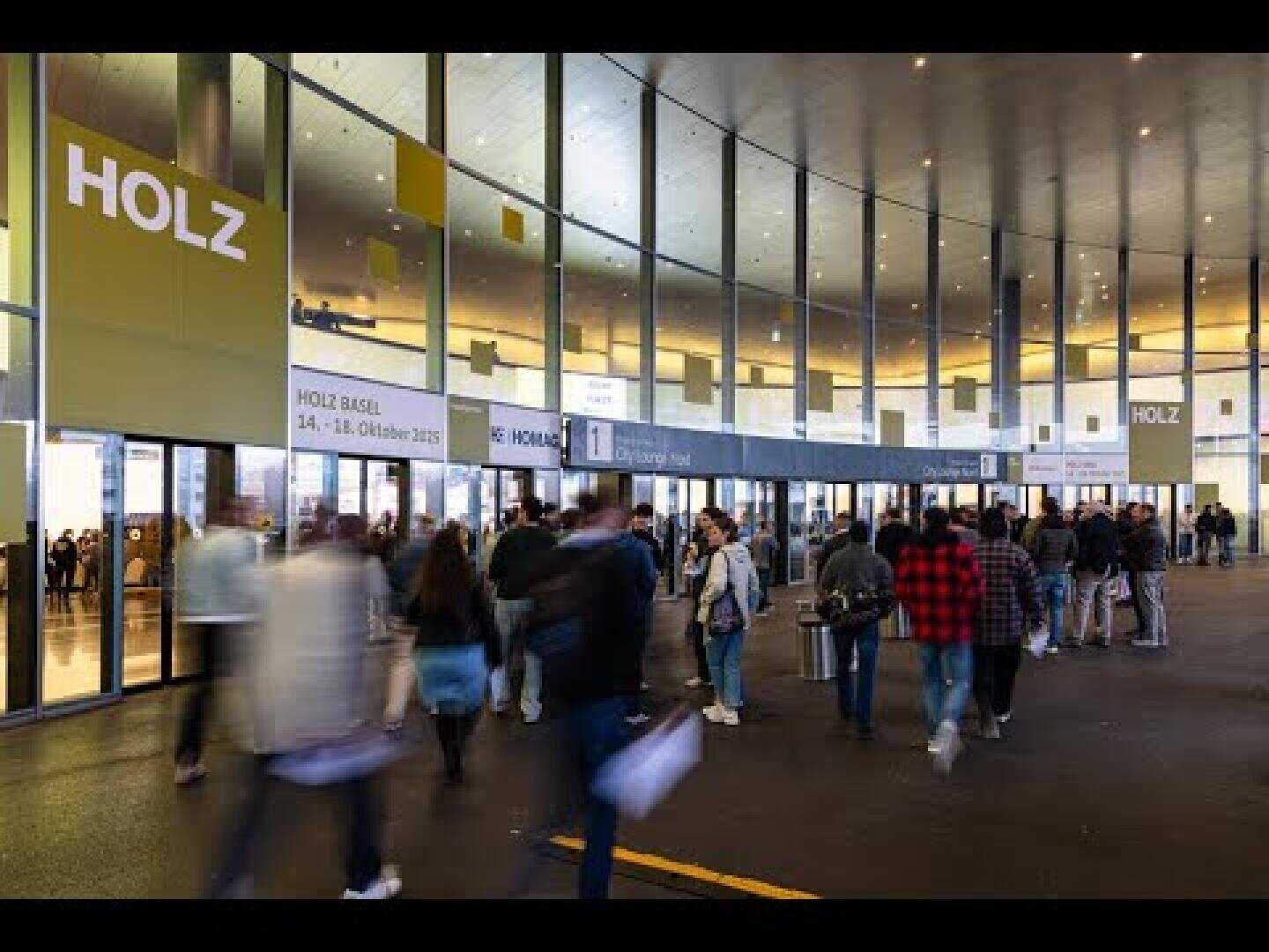 Menschen stehen und gehen vor einem großen, modernen Glasgebäude mit der Aufschrift HOLZ über dem Eingang. Die Szene scheint sich auf einer Veranstaltung oder Ausstellung zu befinden, da sich in den Fenstern Spiegelungen abzeichnen.