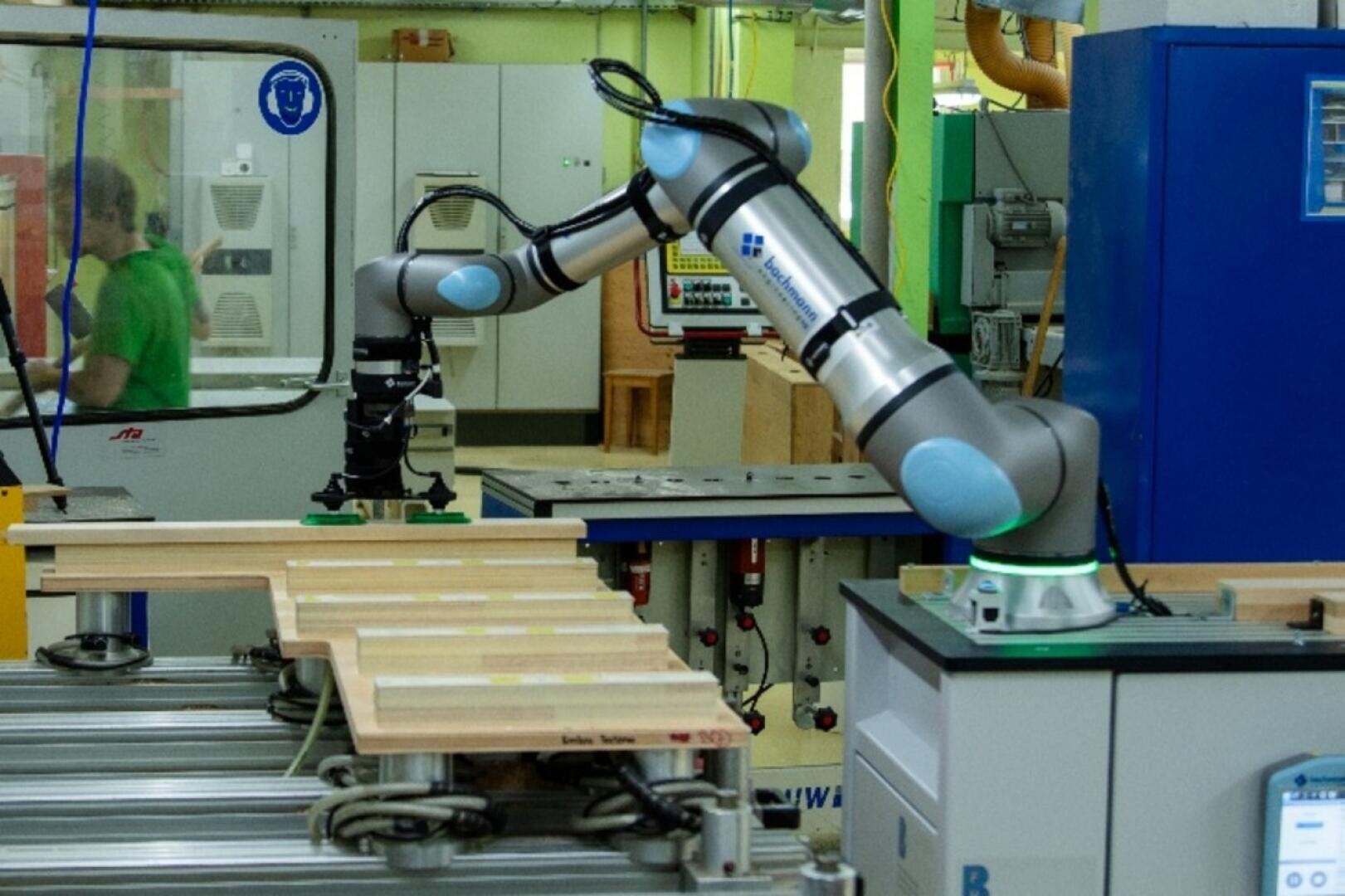 Ein Roboterarm montiert Holzteile auf einem Förderband in einer hell erleuchteten Industriehalle. Im Hintergrund sind Maschinen und eine Person zu sehen.