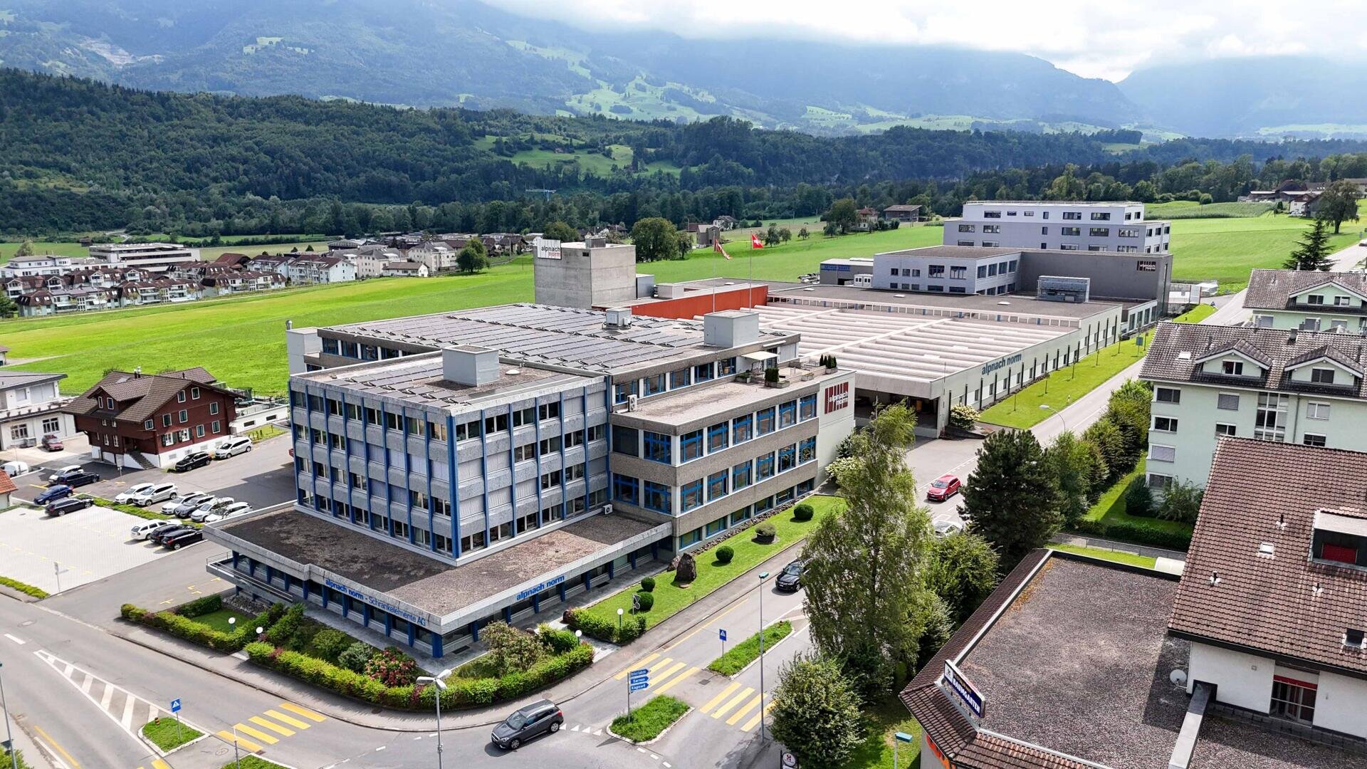 Vue aérienne d'un complexe industriel et de bureaux moderne entouré de champs verts, de bâtiments résidentiels et de montagnes sous un ciel partiellement nuageux. Des passages piétons et des routes bordent la propriété.