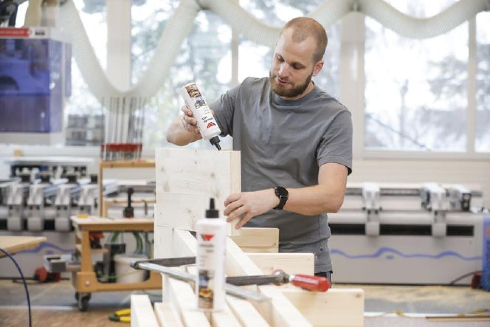 Ein Mann in einem grauen Hemd trägt in einer Werkstatt Holzleim auf ein Holzbrett auf, umgeben von Holzbearbeitungswerkzeugen, darunter ein Hammer und Zwingen auf dem Tisch.