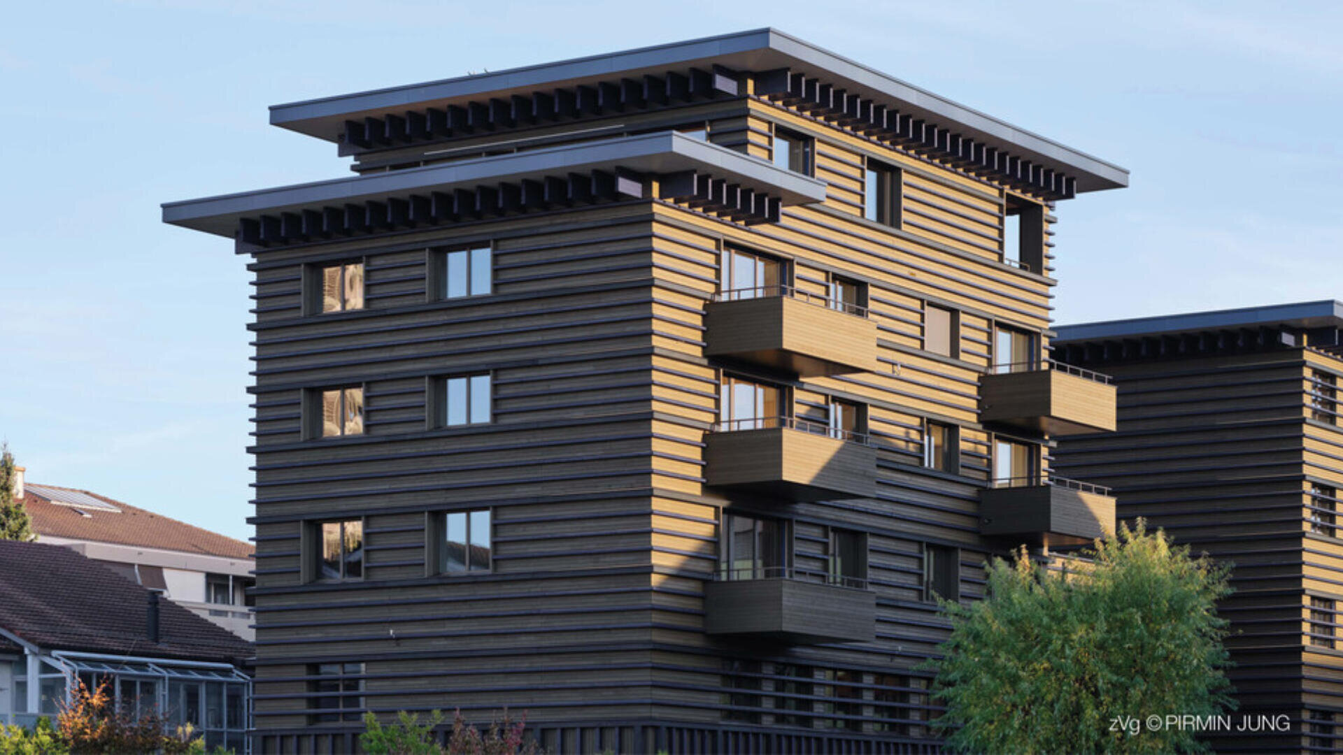 Ein modernes, mehrstöckiges Wohnhaus mit einer gestreiften Holzfassade, vorspringenden Balkonen und großen Fenstern vor einem klaren Himmel mit etwas Grün im Vordergrund.