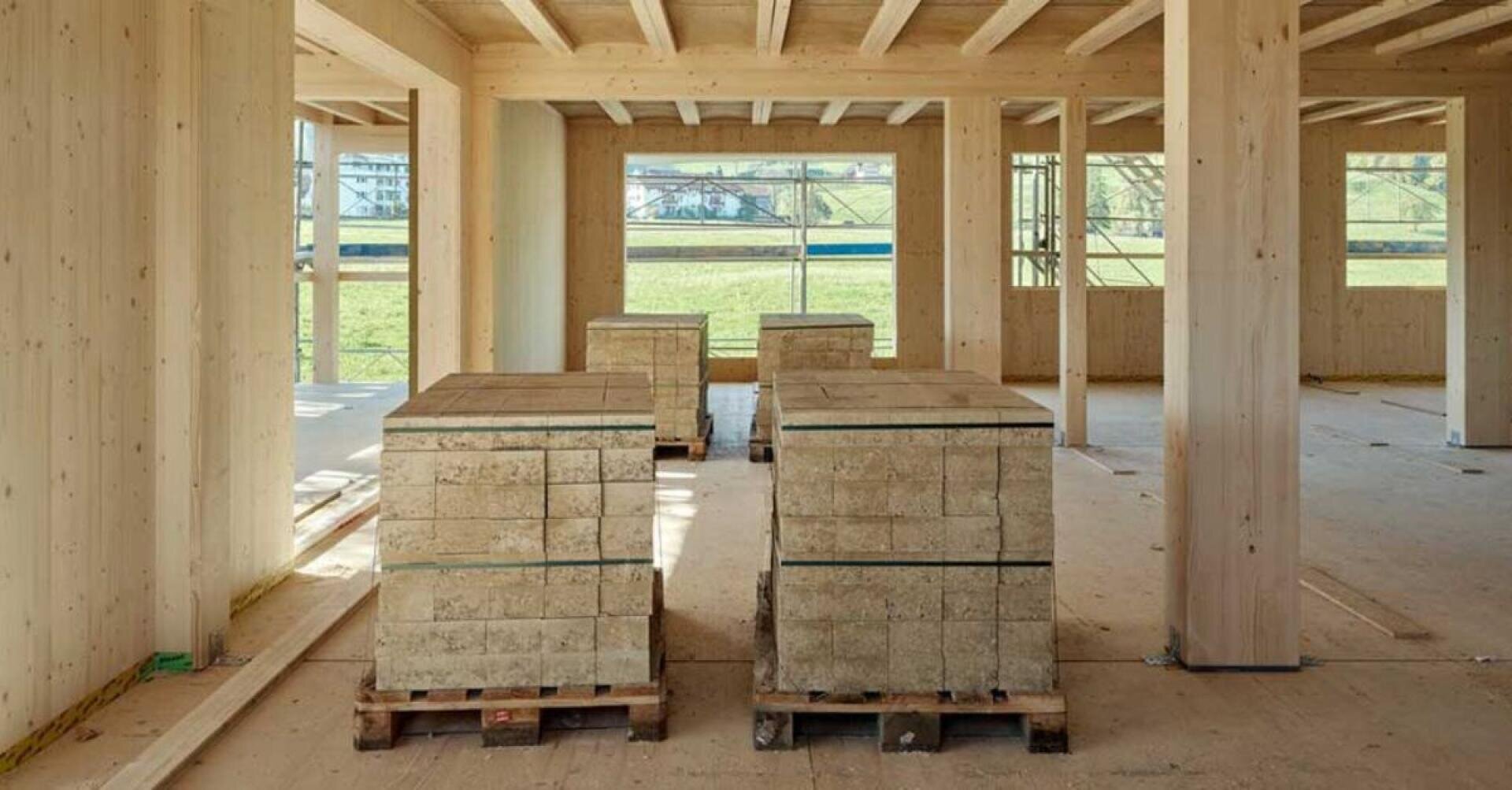 Stapel rechteckiger Bauklötze werden auf Holzpaletten im Inneren eines im Bau befindlichen Holzrahmengebäudes mit großen Fenstern und offenem Blick auf das Gras draußen platziert.