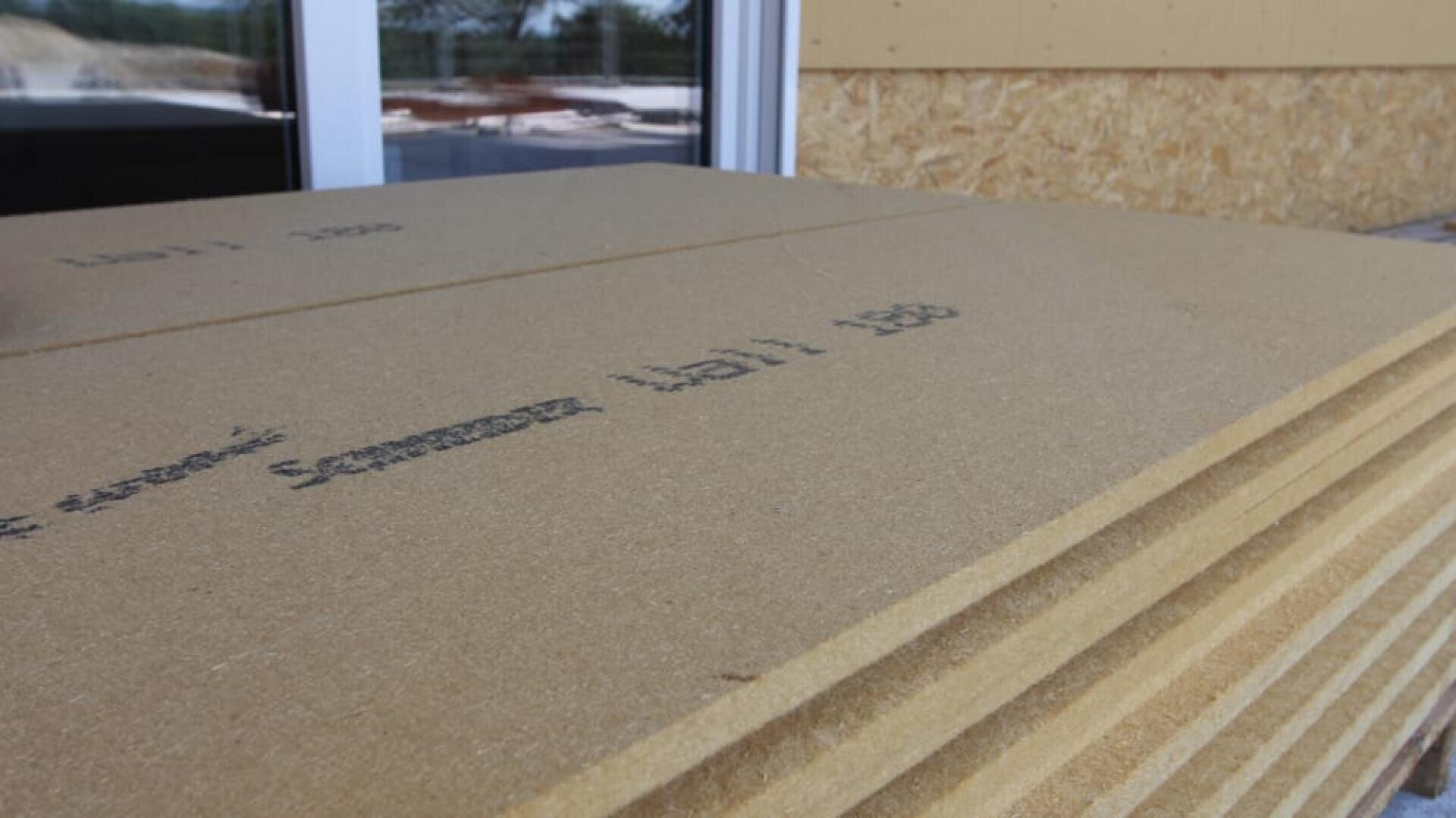 Nahaufnahme gestapelter Faserplatten-Dämmplatten vor einem Gebäude. Im Hintergrund sind teilweise ein Fenster und eine Wand zu sehen. Die Platten weisen aufgedruckte Markierungen auf.