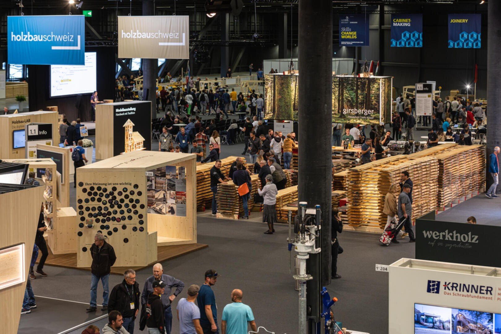 Dans un hall d'exposition très animé, on trouve des constructions en bois, des stands d'information et des expositions sur le thème de la construction en bois. De nombreuses personnes se promènent, explorent les objets exposés et discutent avec les intervenants. De grands panneaux surplombent les différents stands.