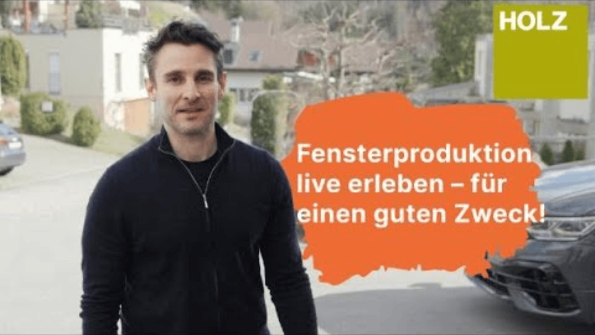 Un homme vêtu d'une veste noire se tient à l'extérieur, dans une rue. À côté de lui, on peut lire en allemand : "Fensterproduktion live erleben - für einen guten Zweck !", avec un logo HOLZ vert dans le coin supérieur droit.