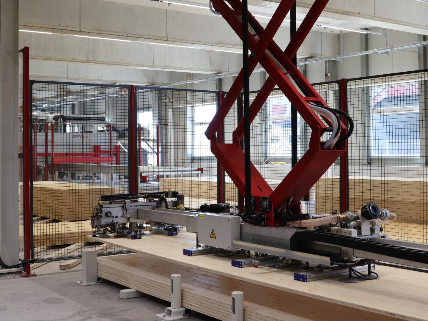 Ein Roboterarm mit roten Halterungen arbeitet in einer modernen Industrieanlage und manipuliert große Holzbretter hinter einem Sicherheitszaun mit Maschinen und Fenstern im Hintergrund.