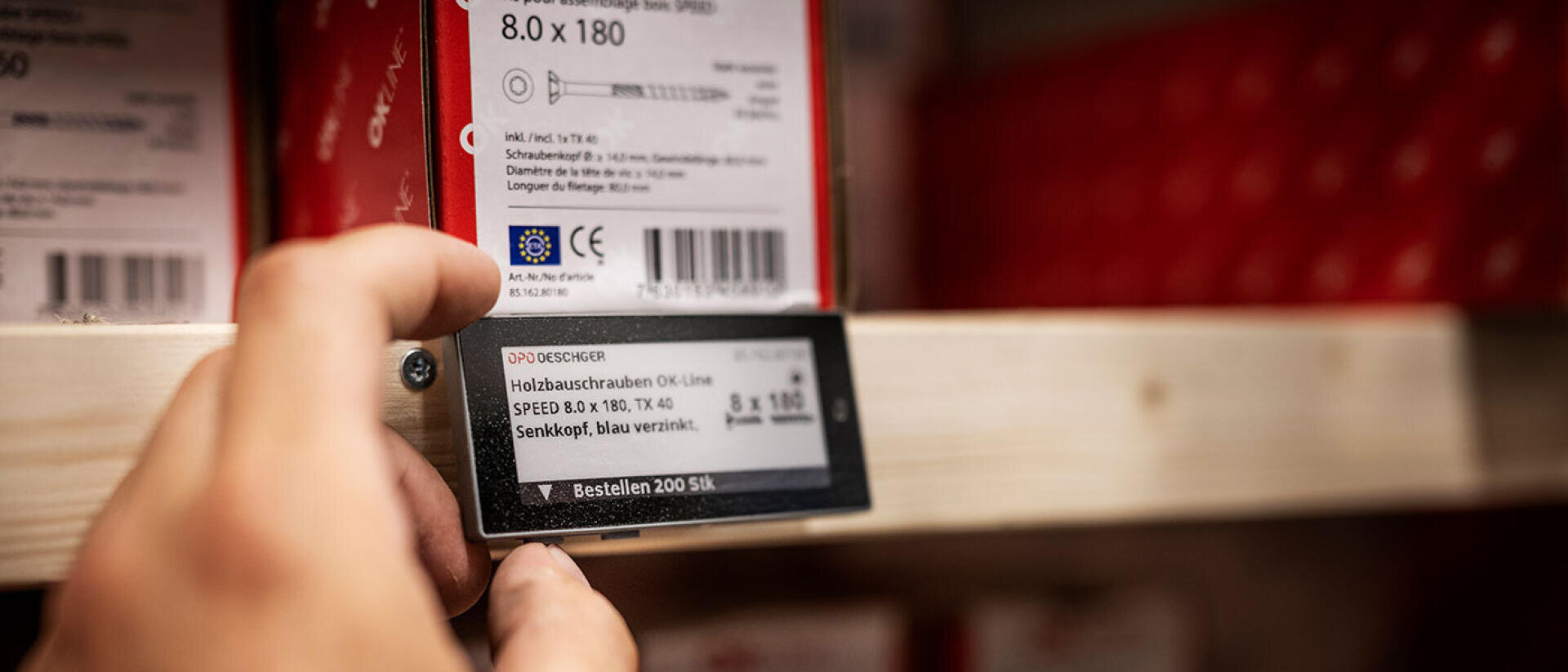 Eine Hand greift nach einem elektronischen Etikett auf einem Regal, das Informationen über Holzschrauben anzeigt, während im Hintergrund Schraubenkästen gelagert werden.