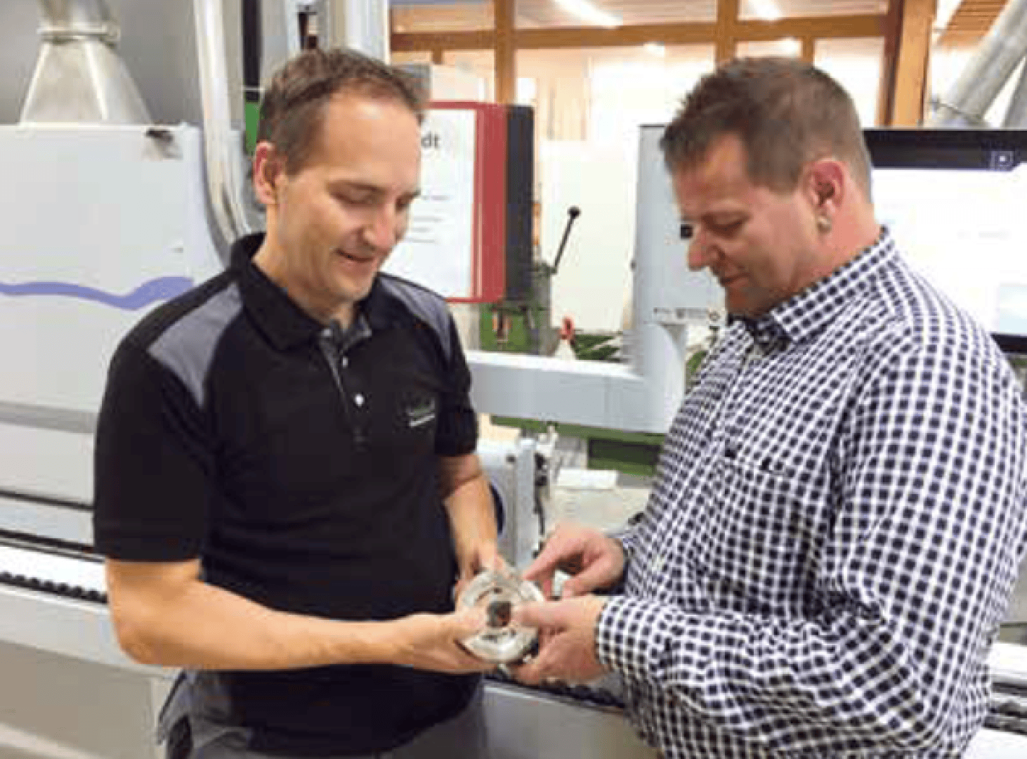 Deux hommes se tiennent dans un atelier et examinent ensemble une pièce métallique. L'un d'eux porte un polo noir, l'autre une chemise à carreaux. En arrière-plan, on voit des machines et des appareils.