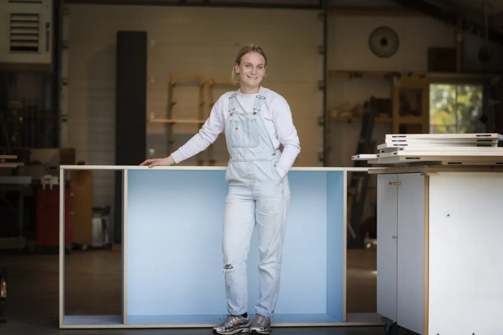 Eine Person in hellblauer Latzhose und Turnschuhen steht in einer Werkstatt und stützt sich auf einen großen, hellblauen, rechteckigen Schrank. Im Hintergrund sind Regale, Werkzeuge und Holzmaterialien zu sehen.