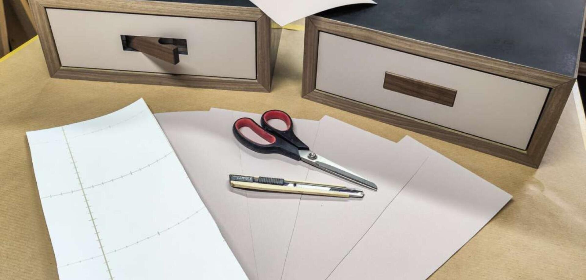 Eine Schere, ein Universalmesser und Papierbögen liegen auf einem Tisch neben zwei Holzschubladen.