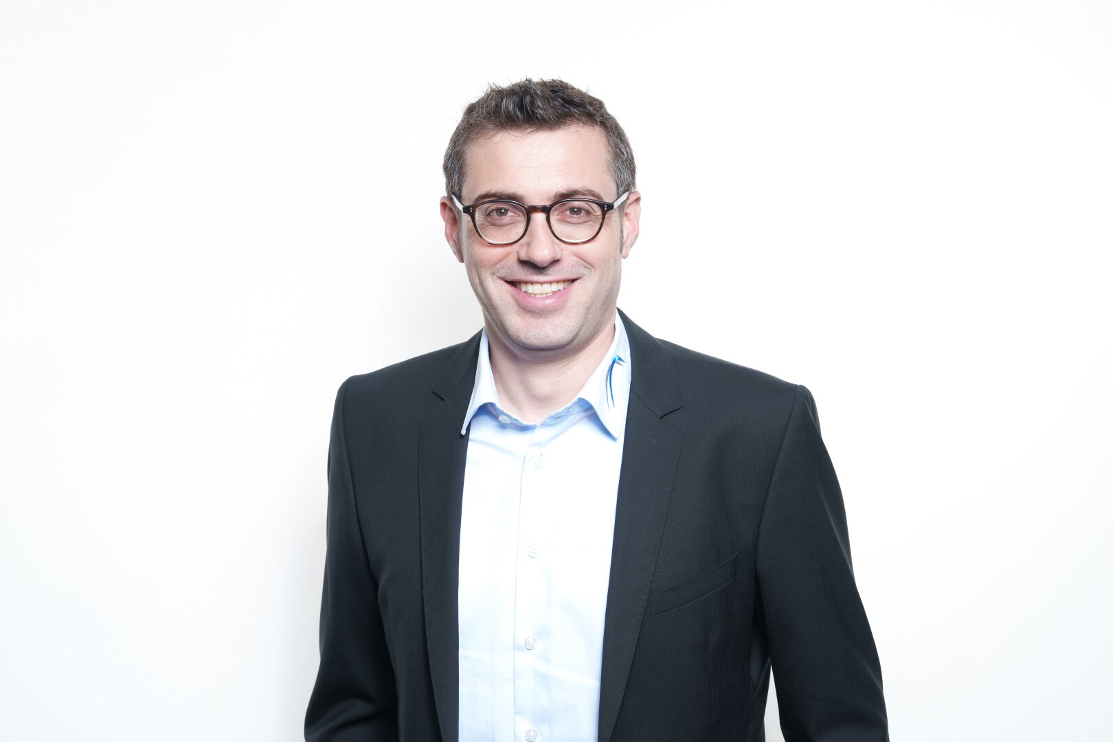 Ein Mann mit kurzen braunen Haaren und Brille, der ein schwarzes Jackett über einem hellblauen Hemd mit Knöpfen trägt, lächelt, während er vor einem schlichten weißen Hintergrund steht.