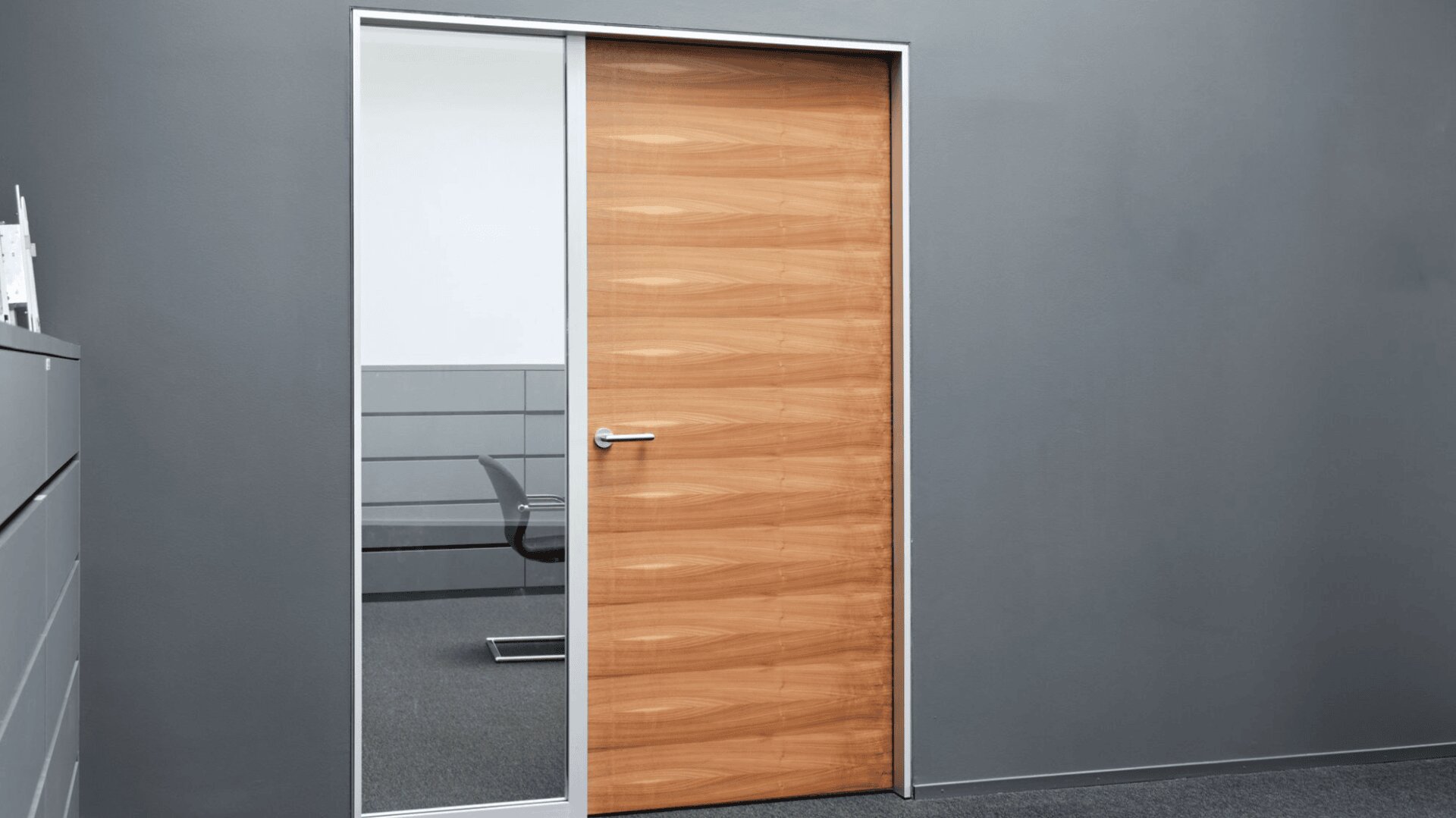 Ein modernes Bürointerieur mit einer geschlossenen Holztür und einer großen Glasscheibe daneben, die einen Teil eines Stuhls und eines Schreibtischs in einem angrenzenden Raum zeigt. Die Wände sind grau und der Boden ist mit Teppich ausgelegt.