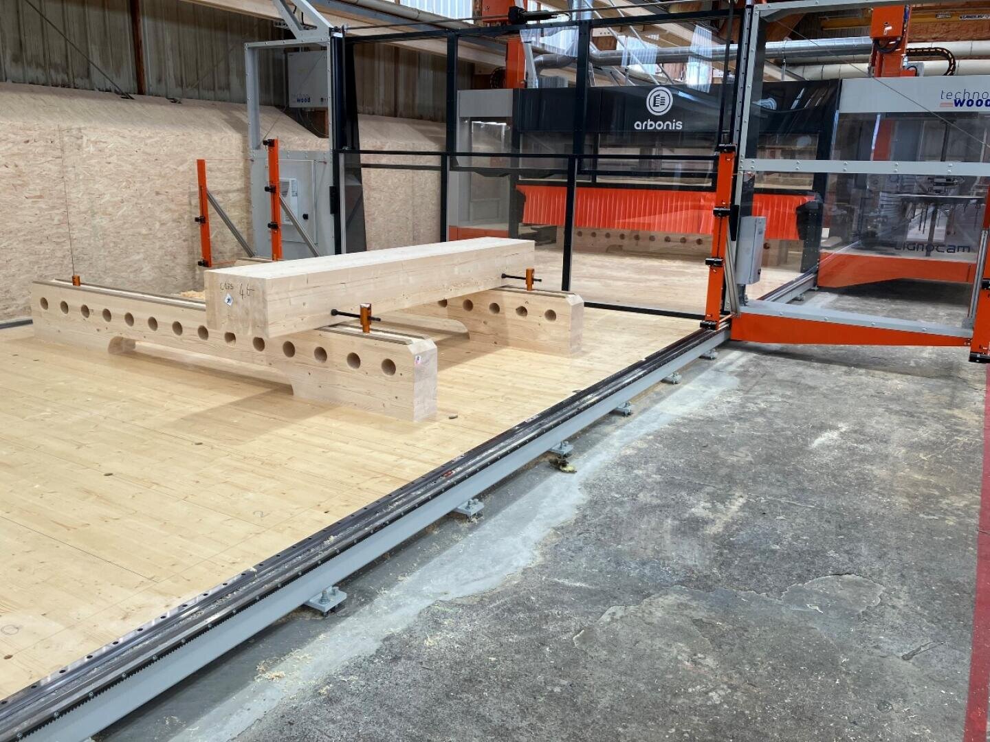 Une grande machine à bois CNC dans un atelier avec une poutre en bois sur la plate-forme de travail, prête pour l'usinage automatique sous un revêtement orange et noir.