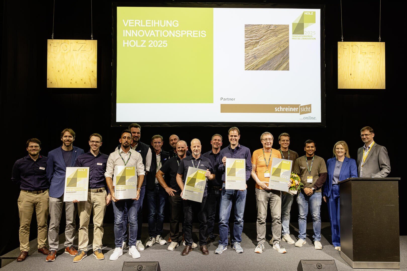 Eine Gruppe von Personen steht auf der Bühne und hält Urkunden und Blumen bei der Verleihung des Innovationspreises HOLZ 2025. Hinter ihnen ist eine große Präsentationsleinwand mit Veranstaltungsdetails zu sehen.