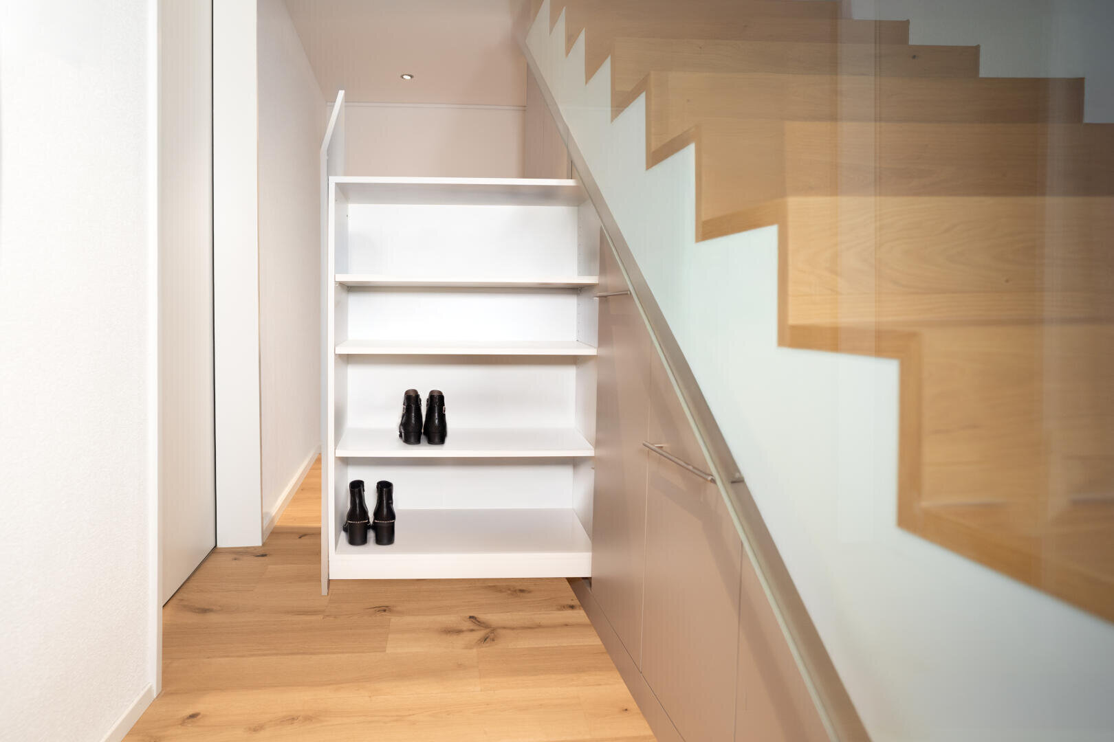 Dans un couloir moderne avec un sol en bois, il y a une étagère blanche coulissante sous l'escalier, dans laquelle se trouvent deux paires de chaussures noires. L'escalier a une rampe en verre.