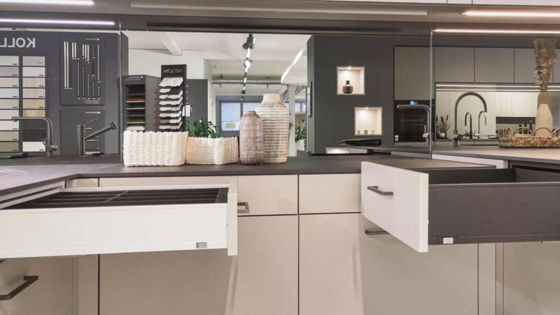 Moderne Küche mit hellen Schränken, zwei offenen Schubladen, dekorativen Körben und Vasen auf der Arbeitsplatte und einem Spiegel, der im Hintergrund weitere Küchenelemente und Geräte reflektiert.