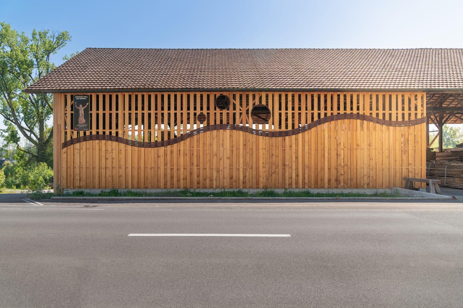 Un bâtiment en bois avec un toit en tuiles, situé à côté d'une rue pavée par une journée ensoleillée, présente un motif ondulé en bois sombre qui s'étend sur le mur extérieur avec une clôture en lattes.