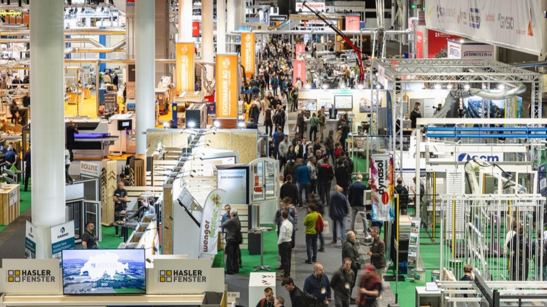Eine belebte Messehalle mit vielen Menschen, die zwischen den Ständen und Auslagen verschiedener Unternehmen umherlaufen, mit Bannern, Schildern und Produktexponaten in einem großen, gut beleuchteten Kongresszentrum.